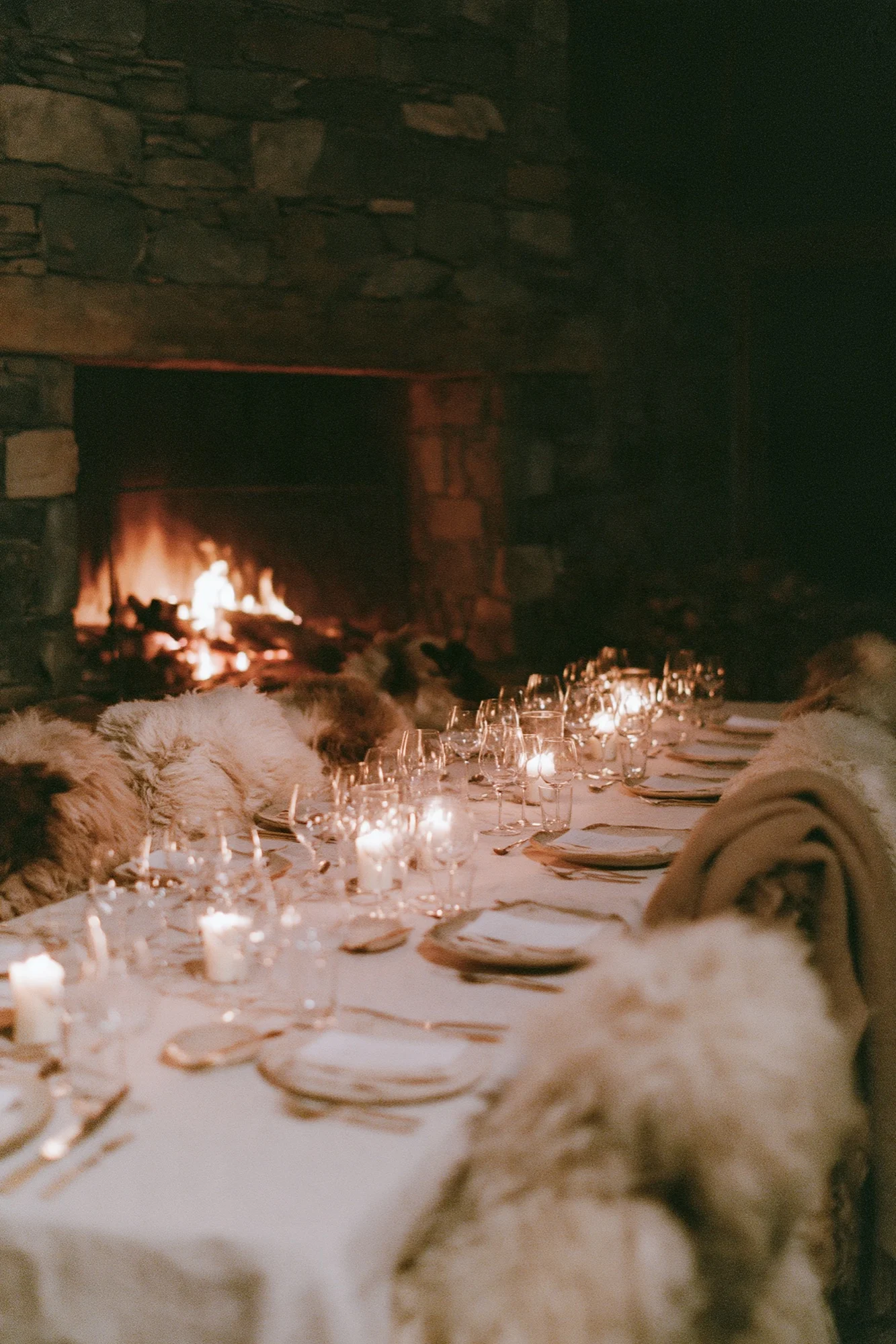 Candlelit long dinner table beside a stone fireplace with sheepskin throws