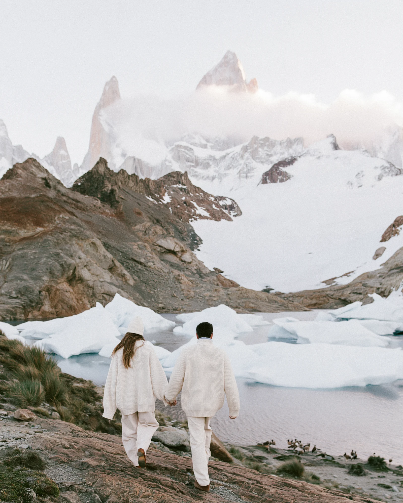 Couple in white walking hand in hand toward snowy Patagonian peaks