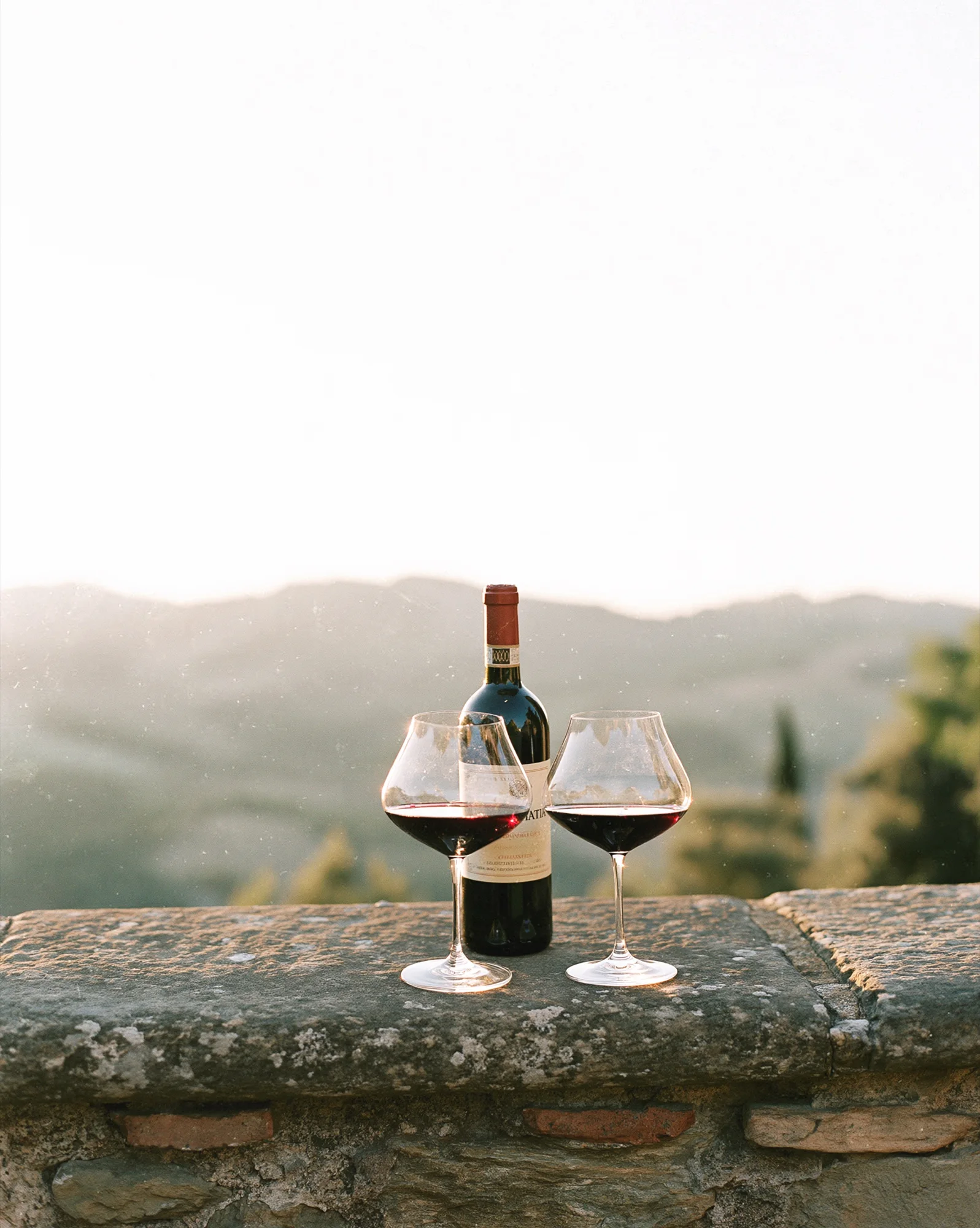 Two glasses of red wine and a bottle on a stone wall above the Tuscan hills